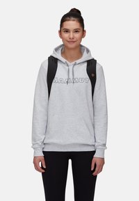 Sweatshirt à capuche gris avec une poche avant et le logo "MAMMUT". Porté avec un legging noir et un sac à dos noir. Texture lisse, coupe décontractée.
