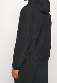 Svart huva jacka, lättviktig, slät textur, elastiska cuffar och nederkant, med en enkel design och inga synliga mönster eller hårdvarudetaljer.