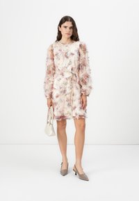 Blommönstrad klänning med genomskinliga, volangärmar, midja som är tajt och en beige bakgrund. Stylad med en liten vit handväska och grå klackar.