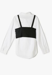 Camisa de manga larga blanca con un superpuesto negro estructurado y tirantes ajustables. La vista desde atrás muestra el cuello y el dobladillo redondeado.