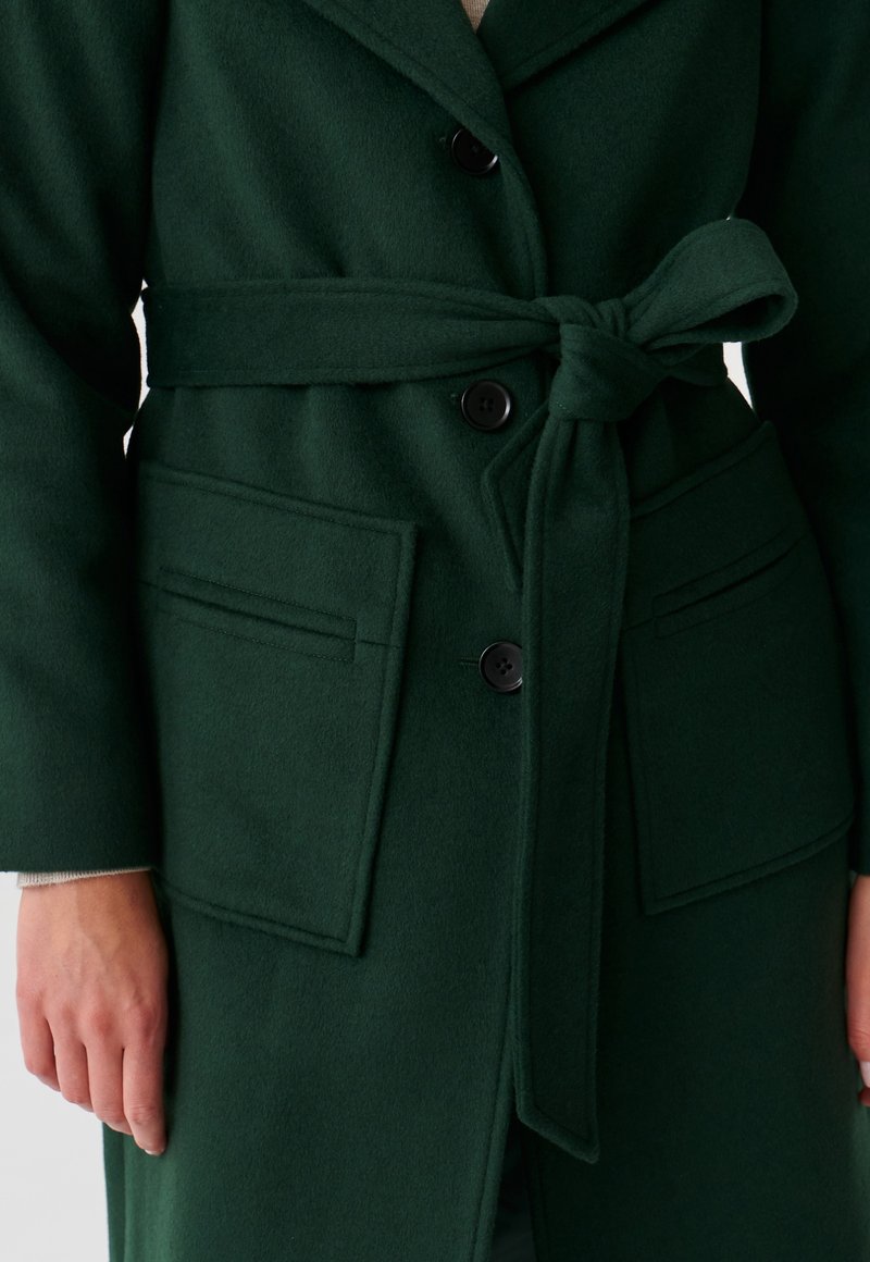 Cappotto in misto lana di colore verde scuro con cintura, colletto a revers e due tasche frontali. Presenta bottoni neri e una texture liscia.