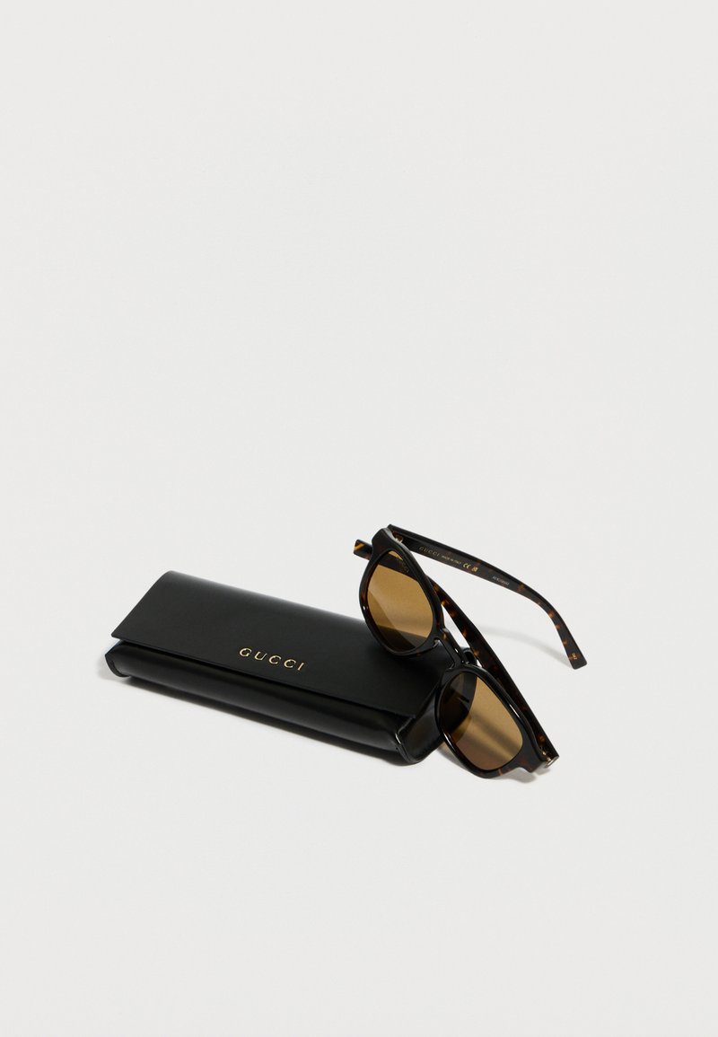 Lunettes de soleil noires Gucci avec des branches en écaille de tortue reposant sur un étui à lunettes en cuir noir Gucci, sur fond blanc.