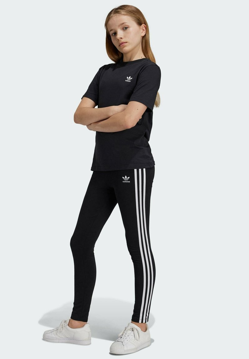 Camiseta negra de Adidas con un logo, combinada con leggins negros que tienen tres rayas blancas. Llevada con zapatillas blancas.