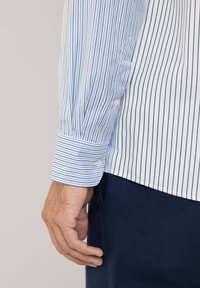 Camisa a rayas con rayas verticales en azul y blanco, material de algodón, puños abotonados, diseño ajustado y textura suave.