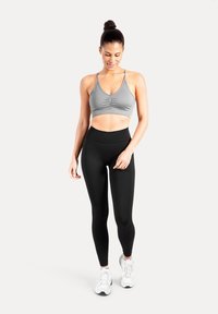 Un soutien-gorge de sport gris avec des bretelles réglables et un détail froncé, associé à un legging noir taille haute, porté avec des chaussures de sport blanches.