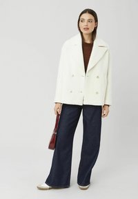 Manteau blanc croisé en tissu texturé, avec de grands revers et des boutons dorés, associé à un jean large foncé et un petit sac à main rouge.