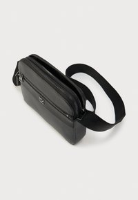 Μαύρη τσάντα χιαστί με υφή στην εξωτερική επιφάνεια, πολλαπλές θήκες με φερμουάρ, ρυθμιζόμενο λουράκι και λεία διακοσμητικά στοιχεία. Συμπαγής και ορθογώνιος σχεδιασμός.