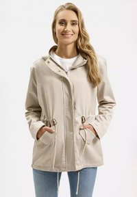 Beżowa lekka kurtka z kapturem, taśmą ściągającą w pasie oraz dwoma przednimi kieszeniami. Gładka tekstura i zapięcie na zamek z metalowymi akcentami.