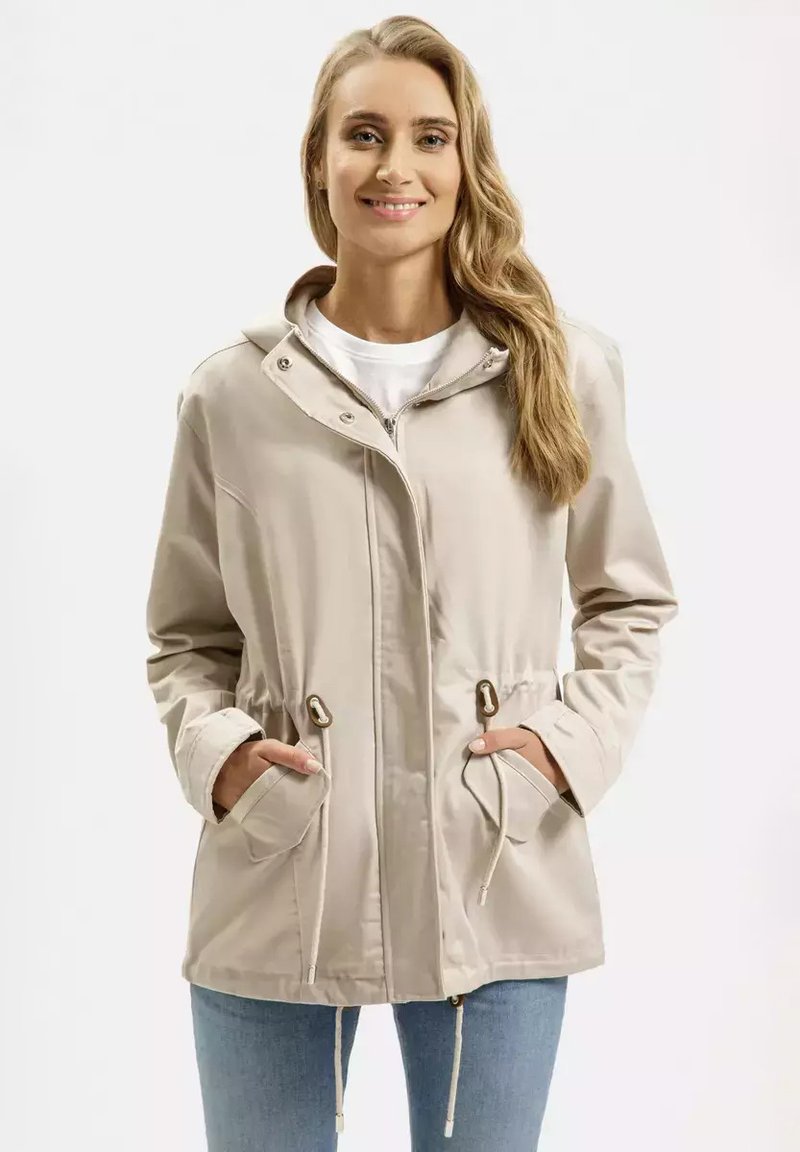 Beżowa lekka kurtka z kapturem, taśmą ściągającą w pasie oraz dwoma przednimi kieszeniami. Gładka tekstura i zapięcie na zamek z metalowymi akcentami.