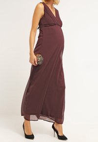 Graviditetsmaxiklänning i djupt burgunder, med V-ringning, plisserad bodice och flödande tyg, parat med en svart geometrisk clutch och klackar.