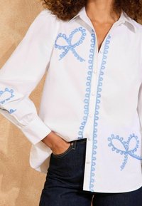 Camicia bianca con ricami a boucle azzurri che formano fiocchi, con maniche a sbuffo e orlo curvo, abbinata a jeans scuri.