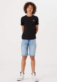 T-shirt nera a maniche corte con un piccolo logo, abbinata a pantaloni corti di jeans blu chiaro. Il modello indossa sneakers alte bianche.