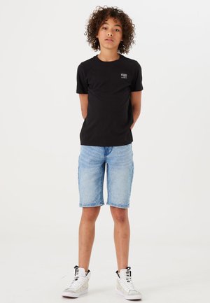 T-shirt nera a maniche corte con un piccolo logo, abbinata a pantaloni corti di jeans blu chiaro. Il modello indossa sneakers alte bianche.