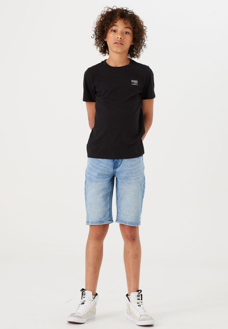 T-shirt nera a maniche corte con un piccolo logo, abbinata a pantaloni corti di jeans blu chiaro. Il modello indossa sneakers alte bianche.