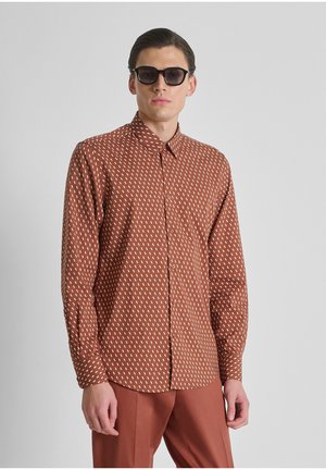 Uomo che indossa una camicia marrone a maniche lunghe con motivo a bottoni, pantaloni marroni abbinati e occhiali da sole neri su uno sfondo grigio semplice.