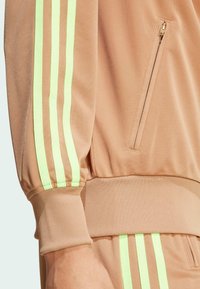 Tanfarbene Sportjacke mit drei neongrünen Streifen an den Ärmeln, einer mit Reißverschluss versehenen Tasche und gerippten Bündchen für einen sicheren Sitz.