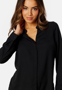 Blusa nera a maniche lunghe con colletto, piastra di bottoni frontale e un texture morbido. Il modello posiziona la mano sul collo.