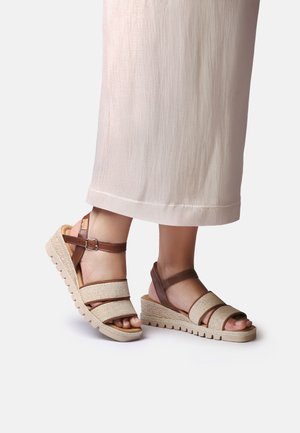 Stopy w beżowych sandałach na koturnie z brązowymi paskami, zestawione z jasnymi szerokimi spodniami na jednolitym tle.