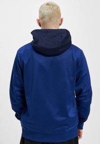 Marinblå hoodie med en mörkare huva, tillverkad av ett slätt tyg. Har en avslappnad passform och ribbade ärmslut. Inga synliga loggor eller mönster.