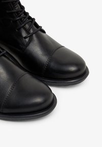 Bottes en cuir noir avec un bout rond et des accents cousus, dotées de lacets noirs et d'une semelle en caoutchouc durable. Texture lisse.