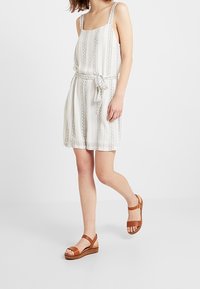 Vit, randig klänning med tunna axelband, ett midjeband och blommönster. Kombinerad med bruna, bandade sandaler på en ljus bakgrund.