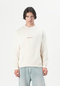 En ung mand med mellemlangt mørkt hår bærer en cremefarvet sweatshirt med et lille orange logo og løse lyseblå jeans foran en ensfarvet baggrund.