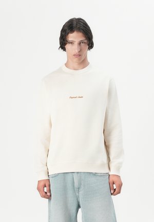 En ung mann med mellomlangt mørkt hår har på seg en kremfarget hettegenser med en liten oransje logo og løse lyseblå jeans mot en ensfarget bakgrunn.