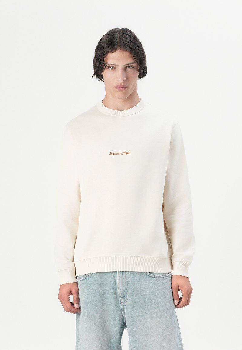 En ung mand med mellemlangt mørkt hår bærer en cremefarvet sweatshirt med et lille orange logo og løse lyseblå jeans foran en ensfarvet baggrund.