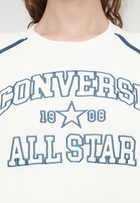Sweatshirt à col ras du cou crème avec texte brodé bleu "CONVERSE ALL STAR", année 1908, et accents bleus aux épaules. Tissu texturé.