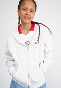 Lys grå hettejakke med glidelås laget av myk stoff, med kengurulomme, snorer og en liten svart Nike-logo på brystet.