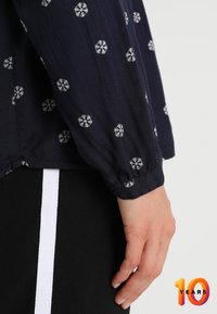 Dunkelblaue Bluse mit weißem Schneeflockenmuster, mit gerafftem Bündchen, teilweise sichtbar auf einem schwarzen Kleidungsstück mit weißem Streifen.