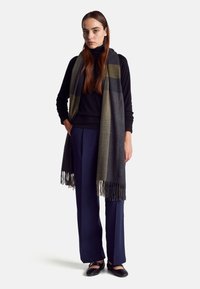 Šedý a olivově pruhovaný šál s třásněmi, kombinovaný s černým rolákem a námořnickými kalhotami, na modelce se rovnými vlasy.