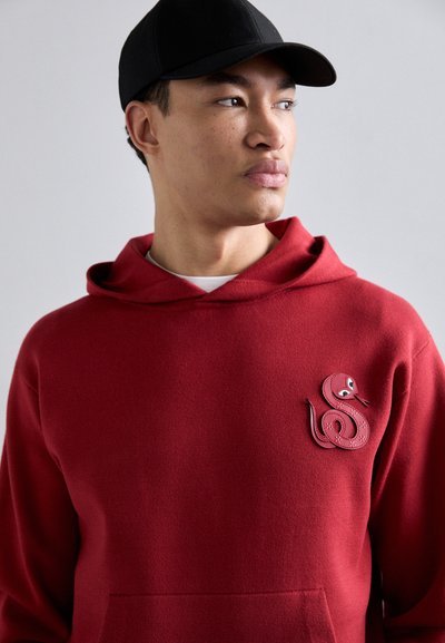 Felpa rossa con una patch di serpente in pelle sul petto, dotata di cappuccio rotondo e tasca frontale. Cappellino da baseball nero indossato in testa.