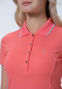 Korálové polo tričko s límečkem, zapínáním na tři knoflíky a stříbrnými detaily na límečku. Obsahuje logo značky na levé části hrudi.