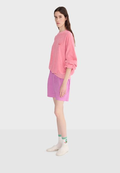 Femme aux longs cheveux bruns portant un sweat-shirt rose ample, un short violet clair, des chaussettes blanches à rayures vertes et des chaussures transparentes, debout de profil.