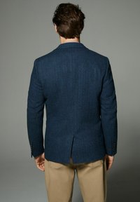 Marineblauer Hahnentritt-Blazer mit einer einzelnen Schlitze hinten, versehen mit einem Spitzrevers und Knopfmanschetten, kombiniert mit beigen Hosen.
