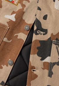 Giacca camouflage con tessuto marrone, beige e nero, caratterizzata da chiusure a pressione in metallo, un rivestimento interno imbottito nero e dettagli di cucitura visibili.
