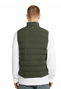 Grön pufferväst med hög krage, med horisontell sömnad och en liten logotyp på baksidan. Bärs över en vit långärmad tröja.