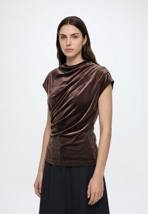 Femme aux longs cheveux foncés portant un haut court en velours marron foncé et une jupe noire, debout devant un fond blanc uni.