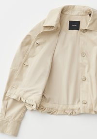 Giacca corta beige con bottoni, colletto, orlo arricciato elastico e fodera interna liscia, stesa in piano e aperta.