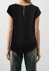 Top nero con maniche corte, collo rotondo e chiusura a zip sul retro. Tessuto liscio, orlo curvo, indossato con jeans blu sbiaditi.