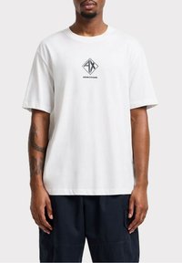T-shirt bianco a maniche corte in cotone con vestibilità ampia. Caratterizzato da un logo centrale nero a forma di diamante con il testo "AX" e "ARMANI EXCHANGE" sottostante.