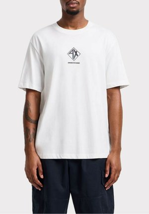 T-shirt bianco a maniche corte in cotone con vestibilità ampia. Caratterizzato da un logo centrale nero a forma di diamante con il testo "AX" e "ARMANI EXCHANGE" sottostante.