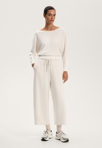 Set de loungewear alb pentru femei, care include un top cu guler larg, drapat și pantalonii cu talie elastică și picioare largi. Purtați cu adidași și șosete de lungime ankle.