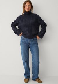 Navy ribbed turtleneck sweater paired with blue straight-leg jeans and brown moccasin-style shoes, featuring relaxed fit and rolled cuffs.