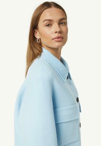 Giacca in lana azzurro chiaro con colletto, grande tasca frontale e bottoni neri. Tessuto morbido e vestibilità leggermente over-size.