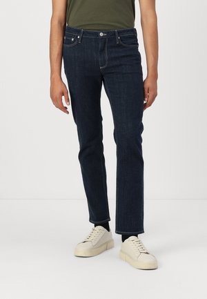Dark blue denim jeans with a straight cut, featuring five pockets, visible stitching, and a button fly. Worn with light-colored sneakers.