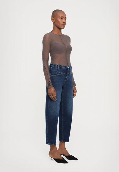 Transparante bruine blouse met lange mouwen en knopen aan de voorkant, gecombineerd met hoge taille blauwe denim jeans; zwarte puntige muiltjes maken de outfit compleet.