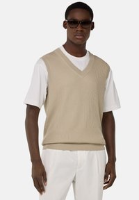 Gilet en tricot beige à côtes avec un col en V, porté sur un t-shirt blanc. Le tissu a une texture lisse, avec un design ajusté et un ourlet droit.