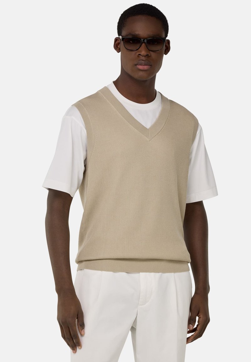 Gilet en tricot beige à côtes avec un col en V, porté sur un t-shirt blanc. Le tissu a une texture lisse, avec un design ajusté et un ourlet droit.